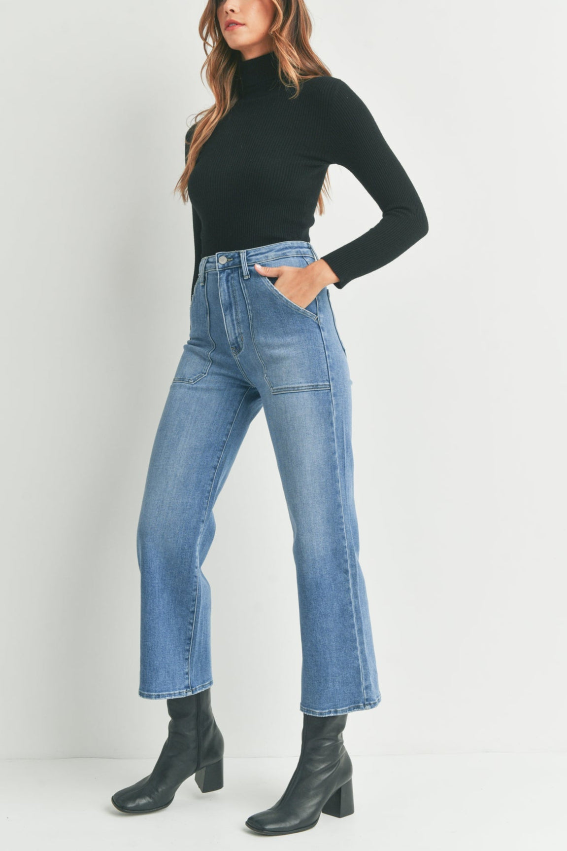 Woman wearing a black turtleneck, blue jeans, and black boots on a white background