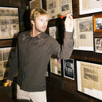Man aiming a dart at framed newspaper clippings on a wall.