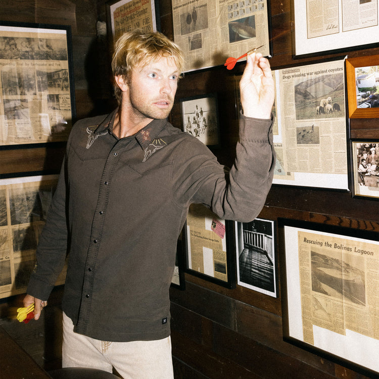Man aiming a dart at framed newspaper clippings on a wall.