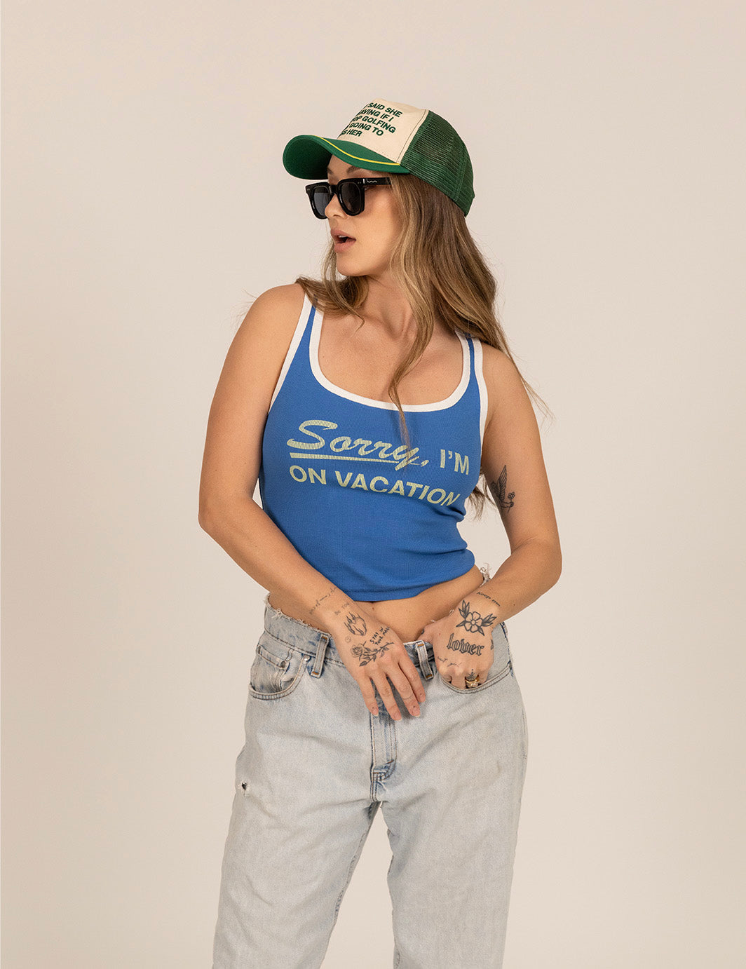 Woman wearing blue ribbed tank top with 'Sorry, I'm On Vacation' text and trucker hat