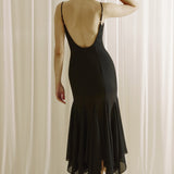 Woman wearing a black dress with a backless design, standing against a white curtain.