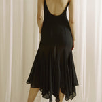 Woman wearing a black dress with thin straps and open back against a white curtain background