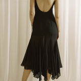 Woman wearing a black dress with thin straps and open back against a white curtain background