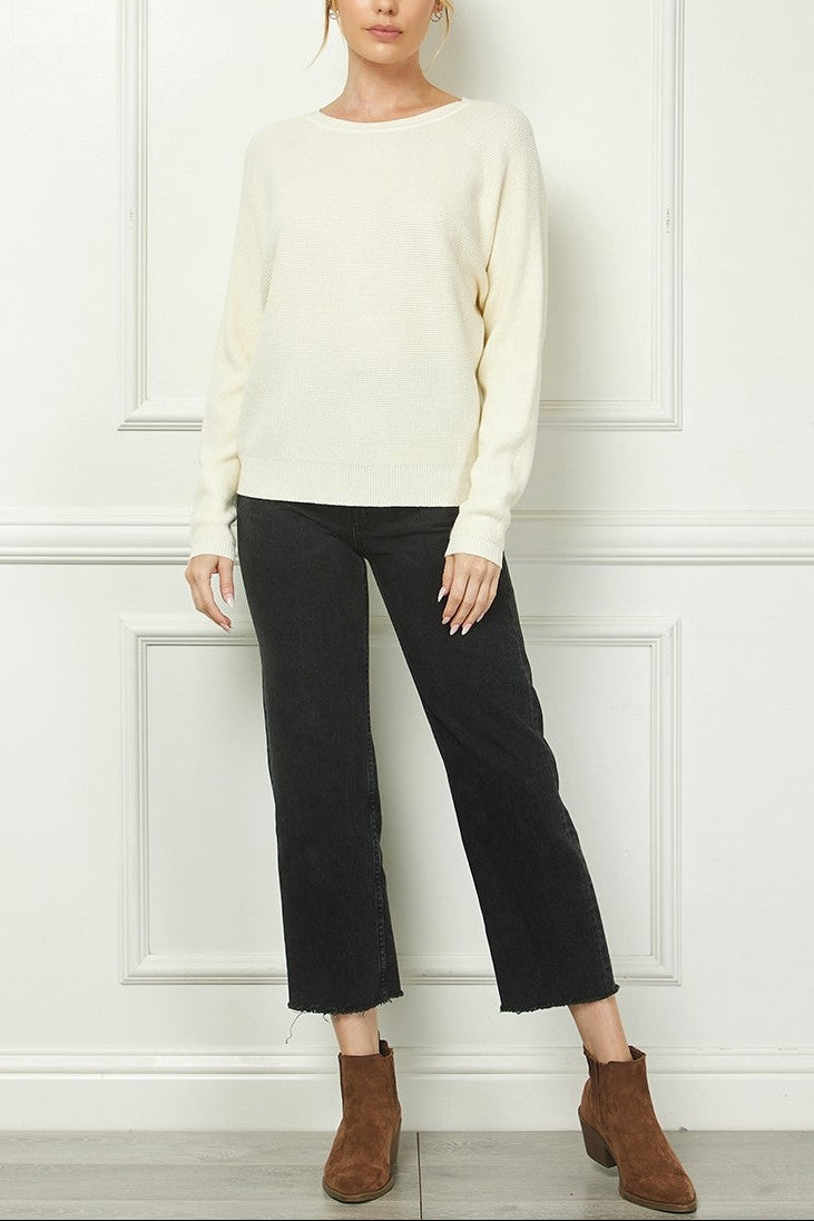 Woman wearing a cream sweater and black pants against a white paneled wall.