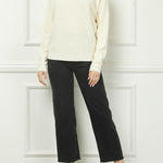 Woman wearing a cream sweater and black pants against a white paneled wall.