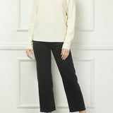 Woman wearing a cream sweater and black pants against a white paneled wall.