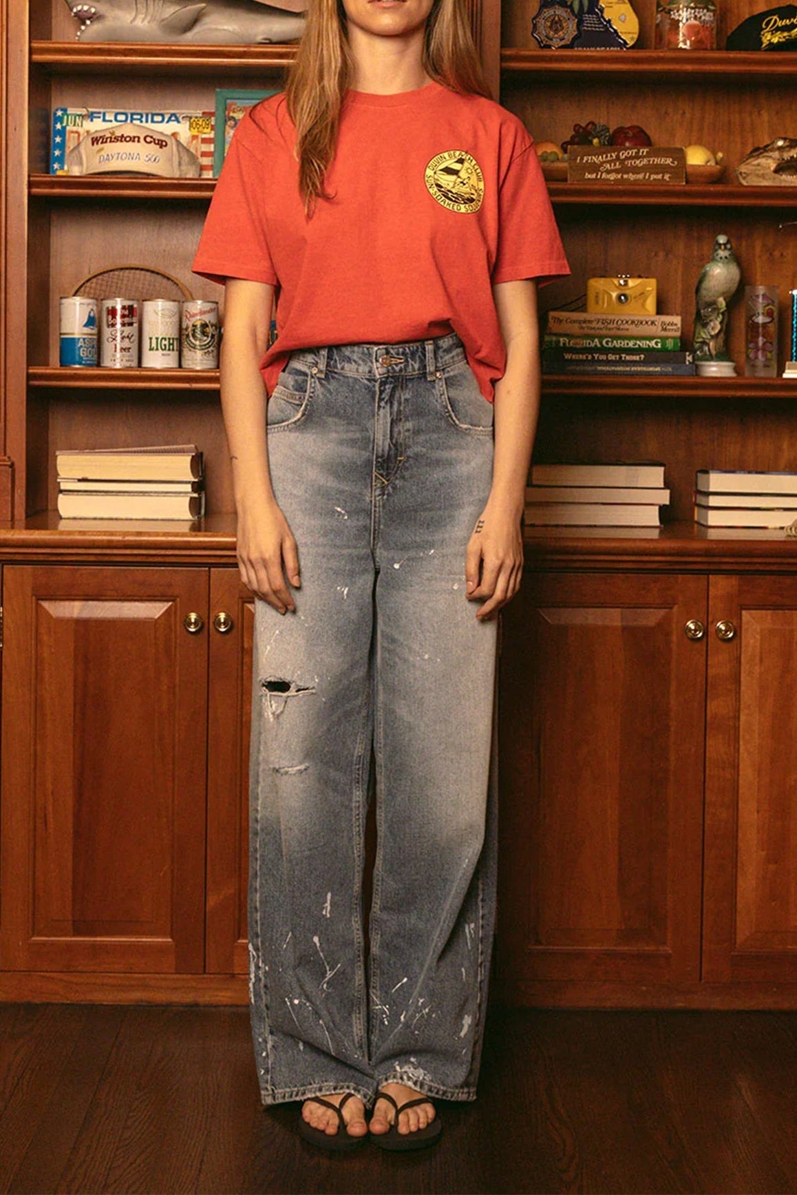 Woman wearing red Duvin souvenir crop tee with distressed jeans standing in front of bookshelf